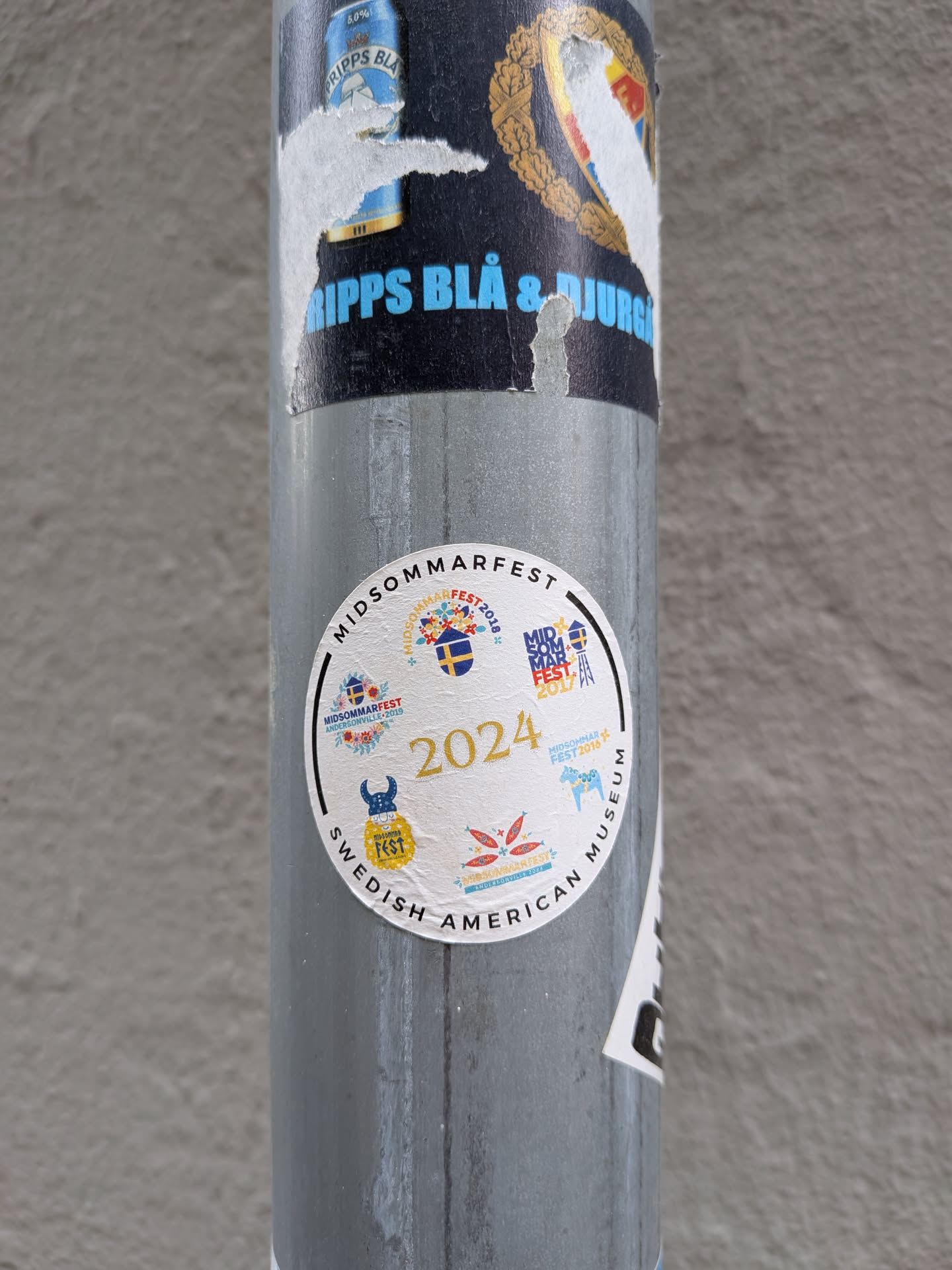 A round white sticker with 'MIDSOMMARFEST' and 'SWEDISH AMERICAN MUSEUM' printed along the border. The center features '2024' in gold, surrounded by smaller logos from past festivals depicting Swedish cultural symbols like a Dala horse, a Viking helmet, and floral motifs.