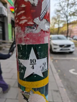A square sticker with a white star on a dark green background. The fraction 10/4 is printed in the center of the star. The sticker is affixed to a pole covered with other stickers and debris.