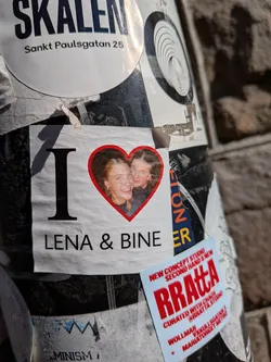 Street sticker The central sticker is rectangular with a white background. It prominently displays a large black capital 'I' on the left. To its right, a vibrant red heart shape frames a photograph of two smiling women, one with blonde hair and the other with darker hair, both looking towards the viewer. Below this 'I' and heart, the names 'LENA & BINE' are written in black capital letters. The sticker appears slightly wrinkled and has a shadow cast across its left side.