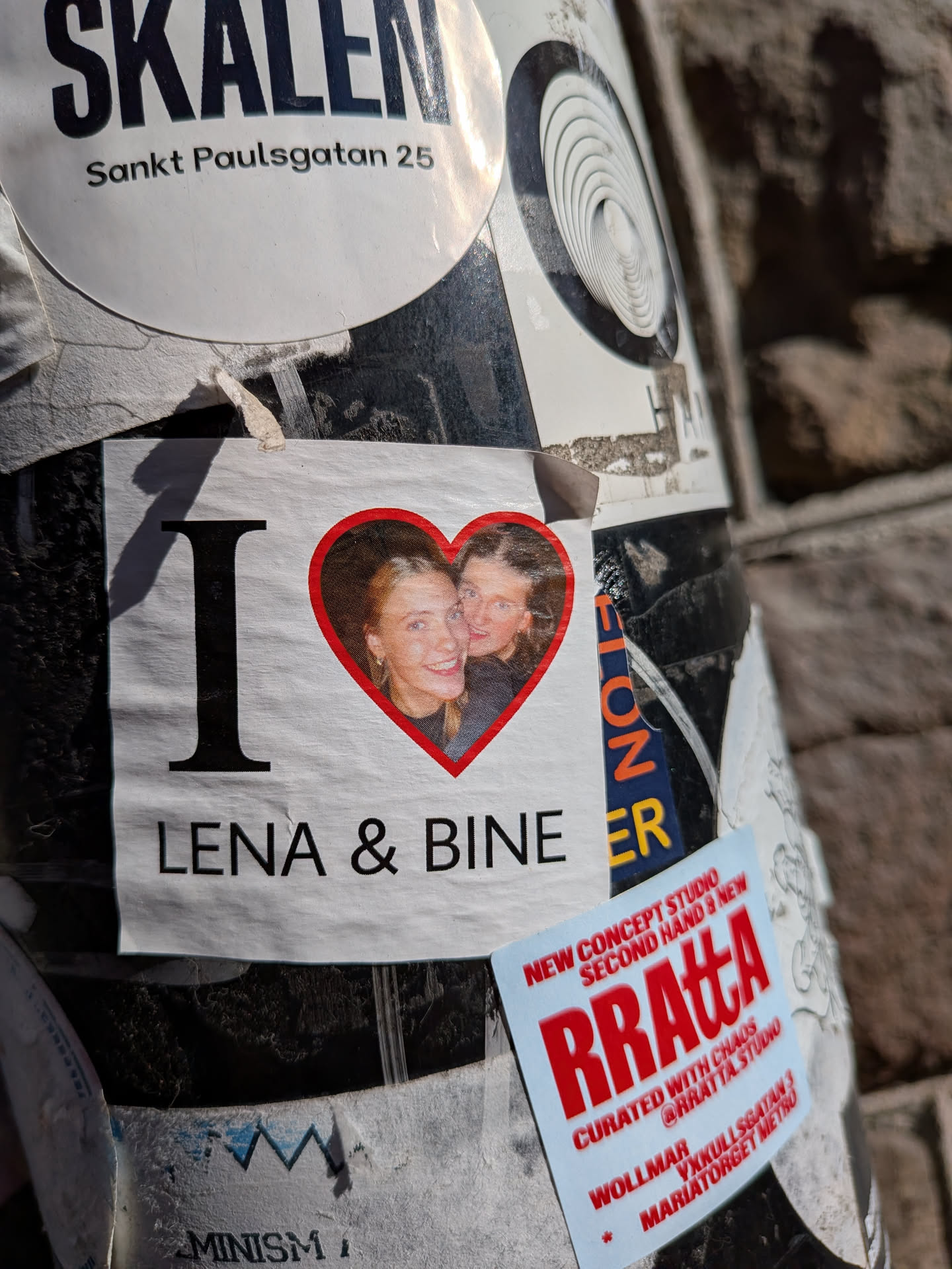 The central sticker is rectangular with a white background. It prominently displays a large black capital 'I' on the left. To its right, a vibrant red heart shape frames a photograph of two smiling women, one with blonde hair and the other with darker hair, both looking towards the viewer. Below this 'I' and heart, the names 'LENA & BINE' are written in black capital letters. The sticker appears slightly wrinkled and has a shadow cast across its left side.