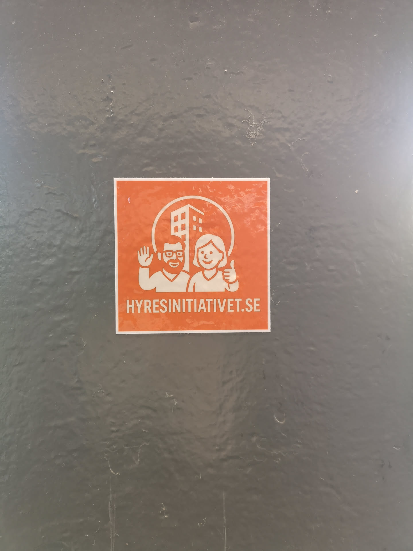 A rectangular sticker with a vibrant orange background and white graphics. The central design features a circular frame enclosing a stylized, multi-story building. To the left of the building, a man with a beard and glasses is depicted waving, while to the right, a smiling woman gives a thumbs-up. Below this central graphic, the website address 'HYRESINITIATIVET.SE' is prominently displayed in white capital letters. The overall style is clean, friendly, and illustrative.