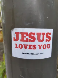 A religious sticker with the message "Jesus Loves You" and a website address WeSabbathKeepers.com. The sticker is white with red lettering and is affixed to a dark green pole.