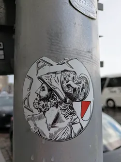 Street sticker The sticker is circular, predominantly white with black linework, resembling an antique engraving or woodcut. It features the profile bust of a bearded man, likely a classical figure such as a Roman or Greek warrior, facing left. He wears an elaborate helmet adorned with intricate, dynamic figures that appear to be in motion or battle. His hair is curly and flows from beneath the helmet, and he has a full, curly beard. Around his shoulder, draped fabric or armor is visible, fastened by a circular brooch. In the upper left portion of the circular design, the stylized capital letters 'PAX' are prominently displayed. To the right of the figure's shoulder, a small shield or crest is depicted, featuring a white field with a red triangular or diagonal section. The overall aesthetic is one of historical gravitas and classical artistry.