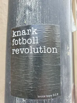 Street sticker A black and white sticker with the text "knark fotboll revolution" and the words "bronx boys D.I.F." at the bottom. The sticker is affixed to a gray pole and shows some wear.