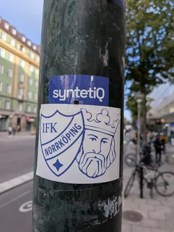 Street sticker A white rectangular sticker with blue illustrations. On the left side, it features the shield of the Swedish sports club IFK Norrköping, containing the text 'IFK NORRKÖPING' and a small four-pointed star at the bottom. On the right side, it depicts the crowned, bearded head of Saint Olof, which is the historical symbol and coat of arms of the city of Norrköping.