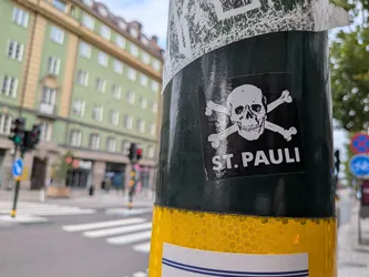 Street sticker A square black sticker featuring a white skull and crossbones illustration, a symbol associated with the German football club FC St. Pauli. Below the skull, the text 'ST. PAULI' is printed in bold, white, sans-serif capital letters. The sticker is affixed to a dark green metal pole with a yellow reflective band visible below it. The background shows a blurred city street with a green building and a pedestrian crossing.