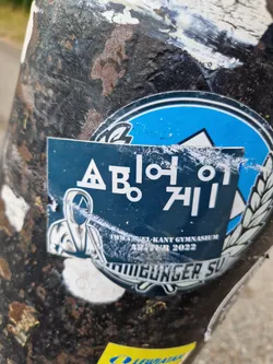 A dark blue sticker with the text "Apollo 711" in a stylized font. There's also an image of a hooded figure and the text "Twiel-Kant Gymnasium Ad-Vent 2022". The sticker appears to be weathered.