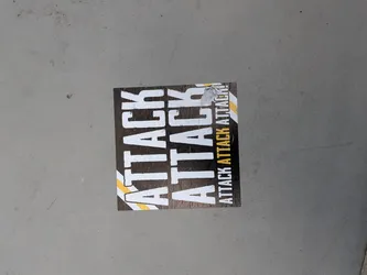 Street sticker A square sticker with a dark, textured background. It displays the word 'ATTACK' twice in large, distressed white font, flanked by diagonal yellow and white stripes. A smaller column on the right repeats 'ATTACK ATTACK ATTACK!' in white and yellow.