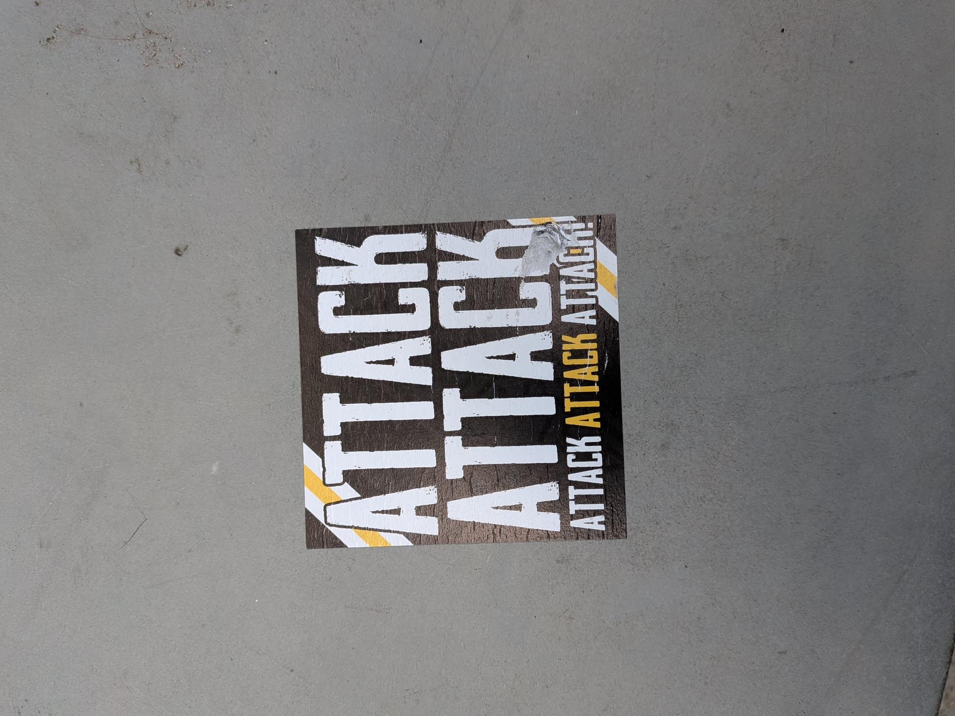 A square sticker with a dark, textured background. It displays the word 'ATTACK' twice in large, distressed white font, flanked by diagonal yellow and white stripes. A smaller column on the right repeats 'ATTACK ATTACK ATTACK!' in white and yellow.