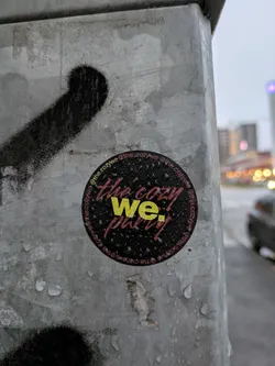 Street sticker Round sticker with the text "The Cozy We Party" in yellow and red on a dark background. The sticker is attached to a gray concrete pole outdoors, and there are water droplets on it. The text @the.cozywe is written around the circle.