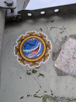 Street sticker A circular die-cut sticker featuring a blue cartoon shark with sunglasses and a wide grin. The character is enclosed in a highly detailed, baroque-style golden frame with repeating cursive text along the inner rim.