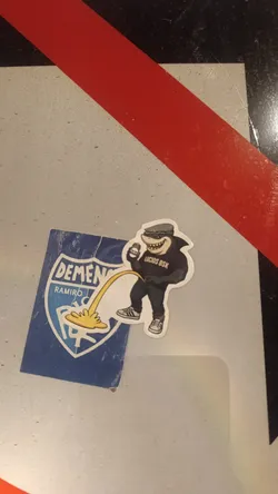 Street sticker The image shows two overlapping stickers on a light grey surface. The background sticker is a blue shield featuring the white text 'DEMENTES' and 'RAMIRO' with a monogram logo. Overlapping this is a die-cut sticker of an anthropomorphic shark character. The shark is wearing a grey patterned flat cap, a black t-shirt with the text 'LUCIOS BSK', dark pants, and sneakers. It holds a beverage can in one hand while urinating a yellow stream onto the blue sticker.