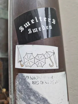 Street sticker A small sticker featuring a simple illustration of three closed umbrellas. The style is minimalist, with black outlines and subtle shading. There is another sticker above this one with text that reads "Swelters Sweden". There are some other unreadable stickers below the umbrella sticker.