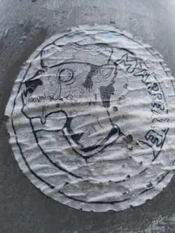 Street sticker A weathered, circular sticker with a monochrome illustration of a creature's head. The style is reminiscent of dark fantasy or gothic art. The sticker shows significant signs of wear and tear, including wrinkles, creases, and some loss of material. Part of the text on the sticker is illegible due to the damage.