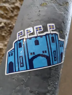Street sticker A vinyl sticker depicting a pixelated blue castle with three small Swedish flags on top.  The art style is reminiscent of 8-bit video games. The sticker is affixed to a grey metal surface.