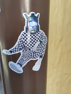This sticker features a unique illustration of a person wearing a checkered jumpsuit and a transformer-like helmet. The style is a mix of streetwear and robotic elements, creating a striking visual contrast. The sticker is likely part of a larger collection or series, given its distinctive design.