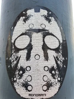 Street sticker A black and white sticker featuring a stylized hockey mask, reminiscent of Jason Voorhees from Friday the 13th. The artwork has a gritty, almost pixelated texture, and is signed "Monomani" at the bottom. It's been placed on a grey metal pole.