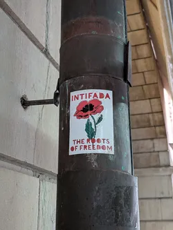 Street sticker The sticker is rectangular with a white background, affixed to a dark, rusty metal pipe. It prominently displays a red poppy flower with a black center and black outlines on its petals. The poppy has a green stem and two green leaves. Above the poppy, in bold red capital letters, is the word "INTIFADA". Below the poppy, also in bold red capital letters, are the words "THE ROOTS OF FREEDOM". The sticker shows some signs of wear, with slightly discolored and peeling edges.