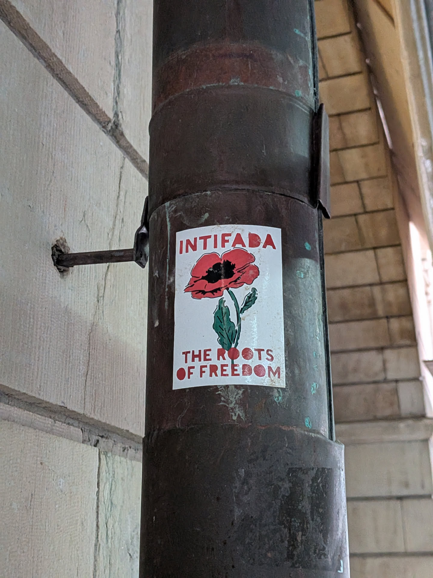 The sticker is rectangular with a white background, affixed to a dark, rusty metal pipe. It prominently displays a red poppy flower with a black center and black outlines on its petals. The poppy has a green stem and two green leaves. Above the poppy, in bold red capital letters, is the word "INTIFADA". Below the poppy, also in bold red capital letters, are the words "THE ROOTS OF FREEDOM". The sticker shows some signs of wear, with slightly discolored and peeling edges.