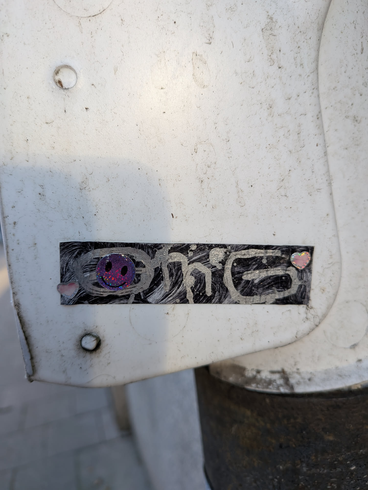 The sticker is rectangular with a black background featuring swirling silver lines. On the left side, there is a small, iridescent pink heart. In the center, a circular, holographic smiley face with sparkling purple and pink tones is prominently displayed. To the right of the smiley face, another small, iridescent pink and yellow heart is visible. The overall impression is one of playful, urban art.