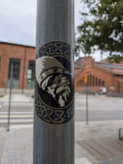 The sticker is circular with a dark blue background. It features a detailed illustration of a Viking's head in profile, rendered in shades of grey and white, wearing a helmet with a crest. The outer edge of the sticker is adorned with intricate knotwork patterns in a lighter blue and gold, reminiscent of Celtic or Norse art. Some text is visible around the circumference, suggesting a connection to Stockholm.