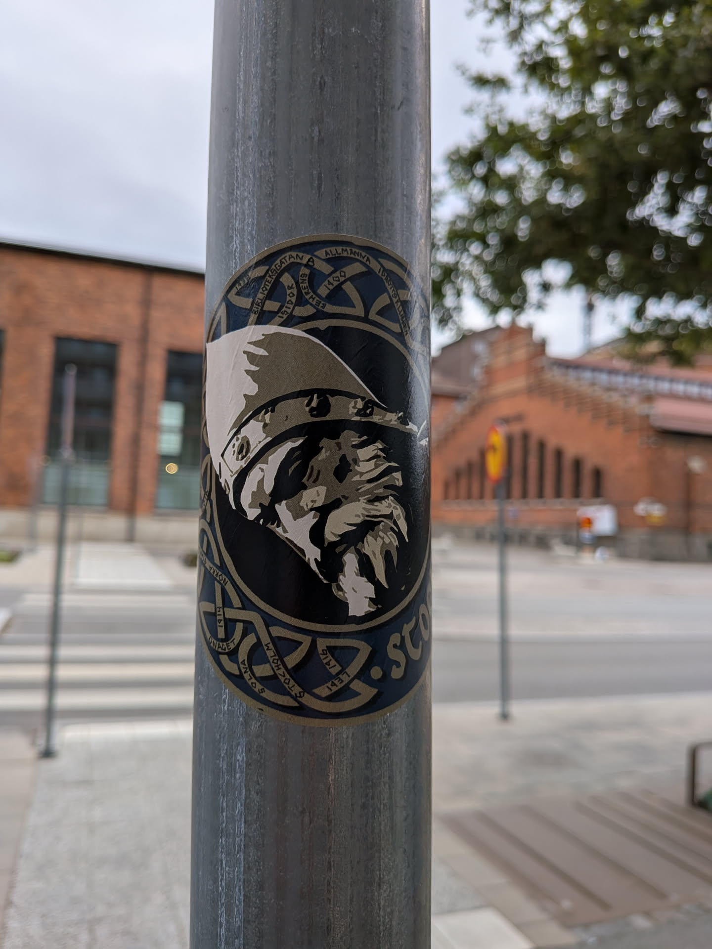 Street sticker The sticker is circular with a dark blue background. It features a detailed illustration of a Viking's head in profile, rendered in shades of grey and white, wearing a helmet with a crest. The outer edge of the sticker is adorned with intricate knotwork patterns in a lighter blue and gold, reminiscent of Celtic or Norse art. Some text is visible around the circumference, suggesting a connection to Stockholm.