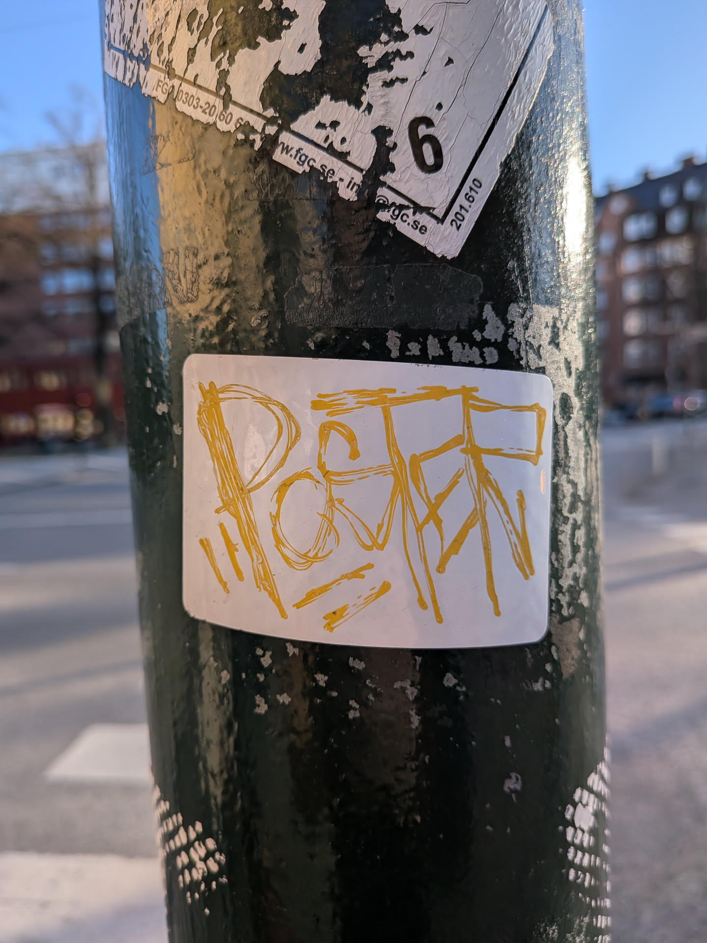 The central sticker is a white rectangle with slightly rounded corners, adhered to a dark, textured pole. It features highly stylized, hand-drawn lettering in bright yellow. The letters appear to form 'POSTR', rendered in a raw, energetic graffiti style with thick, uneven lines. The sticker itself shows minor signs of wear, with some slight peeling at the edges.