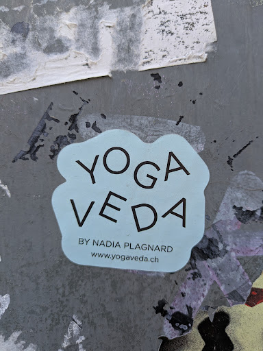 This sticker promotes Yoga Veda by Nadia Plagnard, with a website address provided. The sticker is light blue with dark text, placed on a weathered gray wall with remnants of other stickers.