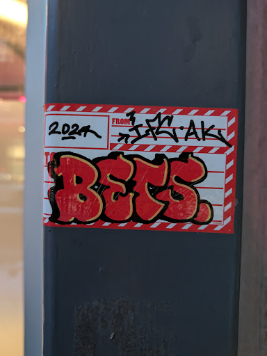 Street sticker A red and yellow sticker with graffiti-style lettering spelling out the word 'BETS'. It's designed to look like a shipping label with '2024' and a tag written above the word 'BETS'. The tag on the top appears to say 'FROM  ...AK'.