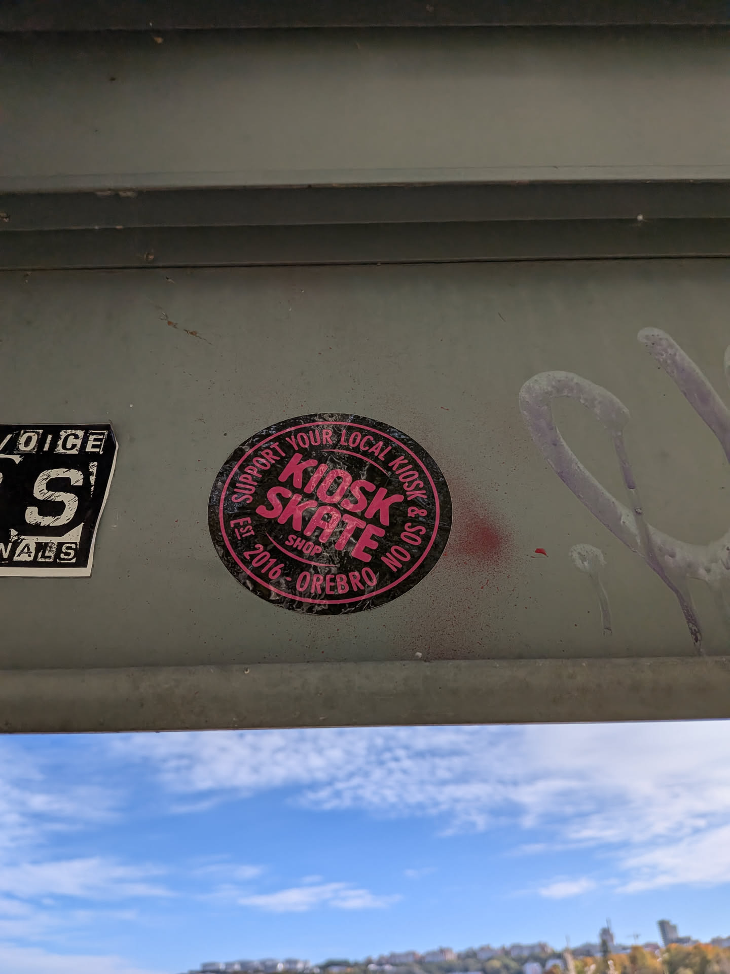 A circular sticker with a black background and bright pink text. The text reads 'SUPPORT YOUR LOCAL KIOSK SKATE SHOP EST 2016 - OREBRO' in a stylized font. The outer edge of the sticker has a pink border. There are also some pink spray paint splatters around the sticker.
