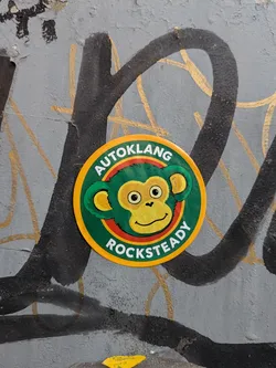 Street sticker A circular sticker with a yellow outer border. The central design features a cartoon monkey head with green fur, a yellow face, and a friendly expression, set against a green background. A thin red ring outlines the monkey's head. Arched along the top in white capital letters is the word 'AUTOKLANG', and along the bottom is the word 'ROCKSTEADY'.