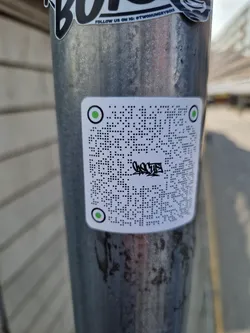 A white square sticker with a QR code in the center and green circles on each corner. There is some black paint on the QR code.  The sticker is affixed to a metal pole.