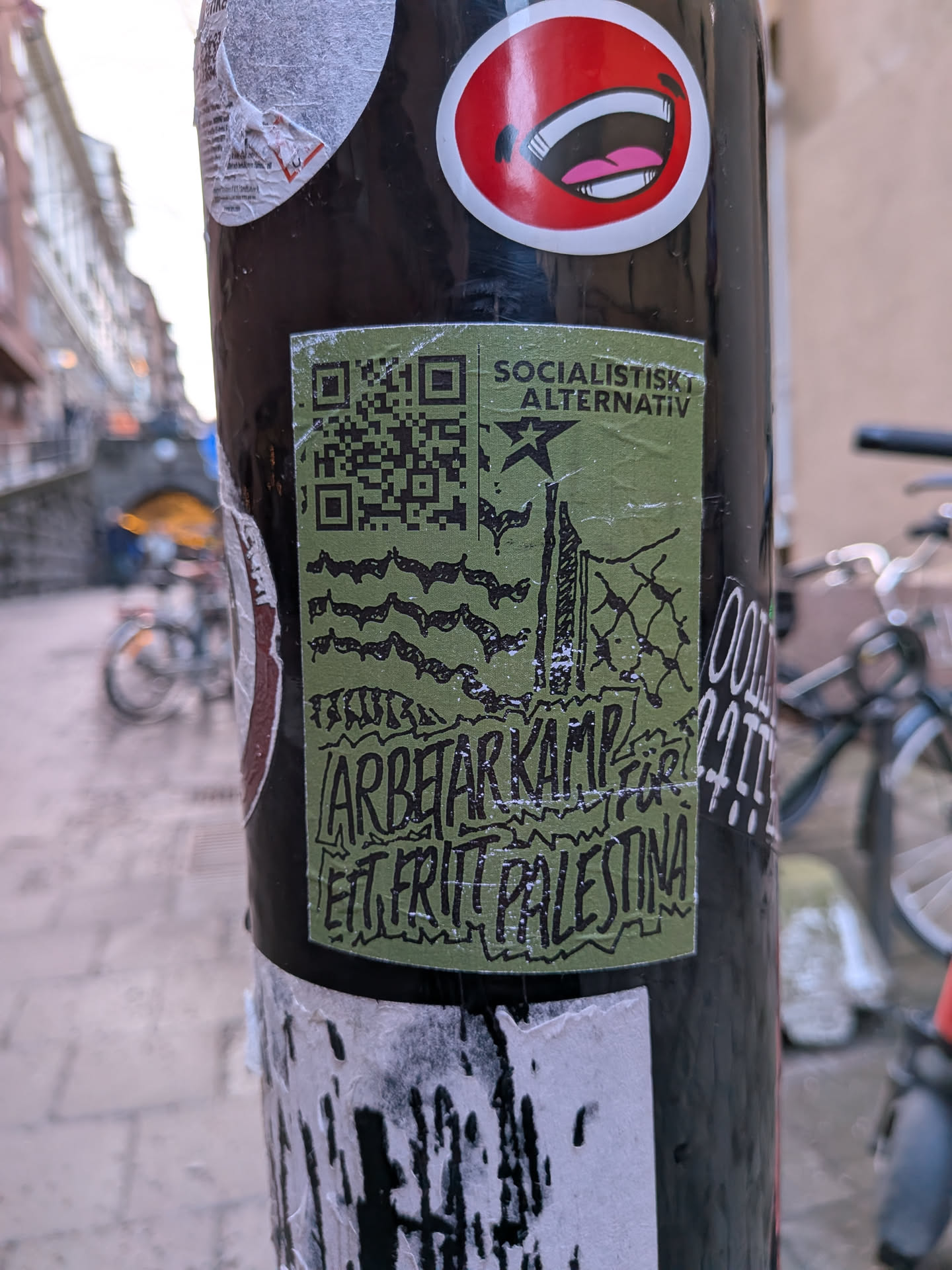 The sticker is rectangular, predominantly olive green with black illustrations and text. In the upper left corner, there's a black QR code. To its right, the text 'SOCIALISTISK ALTERNATIV' is printed in a sans-serif font. Below this text, a black five-pointed star is depicted. The central part of the sticker features a stylized illustration in black, resembling a cityscape or industrial buildings behind a fence, with wavy lines above, possibly representing smoke or a distressed sky. At the bottom, the text 'ARBETARKAMP' is written in a bold, somewhat distressed blocky font, followed by 'ETT FRITT PALESTINA' in a similar, hand-drawn style, with some letters appearing to be outlined or filled with a rough texture. The sticker shows signs of wear, with some white scuffs and minor peeling, indicating it's a physical sticker on a pole.