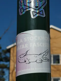 Street sticker The sticker is rectangular, wrapped around a dark green pole, featuring a light purple or off-white background. In the center, there is a black outline drawing of a crocodile or alligator with its mouth wide open, showing teeth. Above the crocodile, in bold white capital letters, is the text "HELA STOCKHOLM HATAR FASCISTER". Below the crocodile, in smaller white capital letters, is "INGA RASISTER PÅ STOCKHOLMS GATOR". The sticker shows some signs of wear and slight peeling at its top edge.
