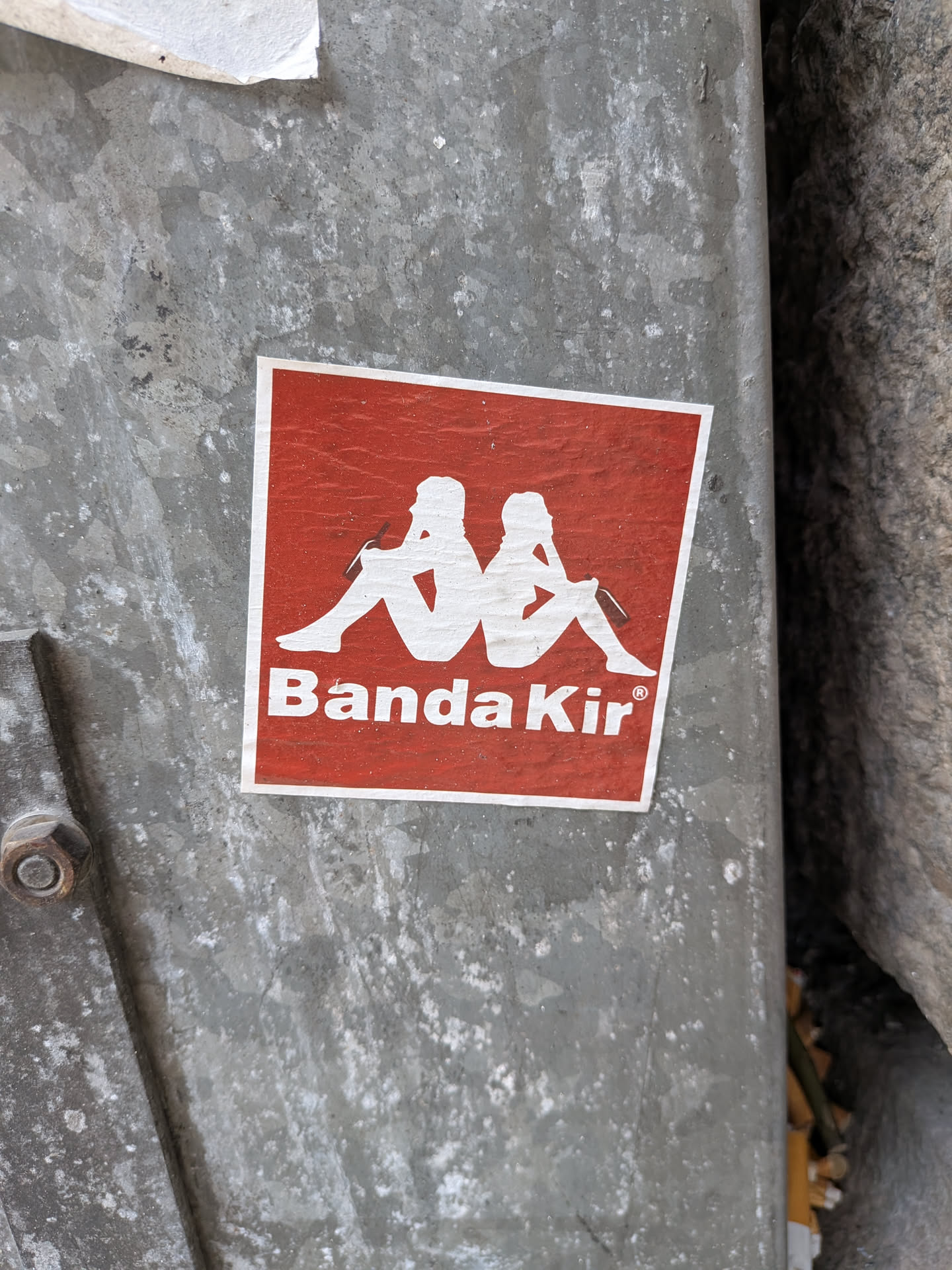 Square red sticker with a white border. Features a white silhouette parody of the Kappa logo, depicting two figures sitting back-to-back holding bottles, above the white text 'Banda Kir®'.