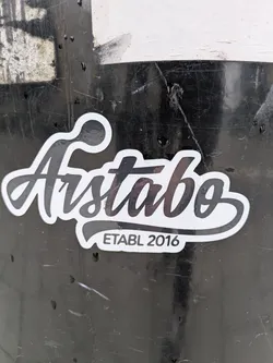 Street sticker The sticker features a prominent, stylized word "Aistabo" in a flowing, elegant script font. It is primarily black with a clean white outline, creating a strong contrast against the dark, scratched background it's affixed to. Below the main text, in a smaller, sans-serif font, are the words "ETABL 2016" on a small, curved banner-like shape, also in black text on a white background with a black outline. The overall shape of the sticker follows the contours of the text.