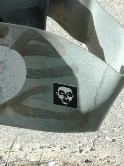 A square-shaped sticker featuring a high-contrast black and white design. The central image is a stylized, ghoulish white face or skull with prominent cheekbones, sunken eyes, and a wide, open mouth revealing teeth. The face is set against a solid black background. The overall style is stark and graphic, resembling a stencil or a simplified illustration.