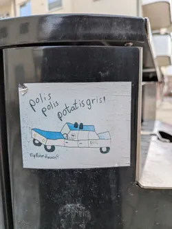 Street sticker A rectangular, hand-drawn sticker featuring a cartoonish police car on a white background. The car is outlined in black, with light blue details on the roof lights and front section, and yellow headlights. A small, simple figure with a sad expression is visible in the driver's seat. Above the car, the text 'Polis polis potatis gris!' is written in black, and below, 'Filip Rahim Hansson' with a small smiley face. The sticker shows signs of wear, particularly a slight tear on the top-left corner, and is affixed to a dark, textured surface.