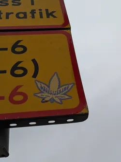 Street sticker The sticker is shaped like a cannabis leaf and is predominantly blue with yellow lettering. The text 'LEGALISERING' is prominently displayed in a stylized font across the leaf. Below this, in smaller text, it reads 'än enda lösningen'. The sticker has a slightly glossy finish and appears to be adhered to a yellow traffic sign with red borders.