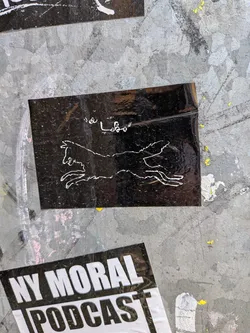 Street sticker The sticker is a rectangular, glossy black background with a white outline drawing of a running animal, resembling a wolf or a wild dog, positioned centrally. The animal is depicted in mid-stride, with its head facing left and its mouth slightly open, showing teeth. Above the animal, in the upper left corner, white handwritten-style text reads 'la Lobo'. The sticker shows some wear, reflections, and minor creases, indicating it's affixed to a textured, possibly metallic, surface.