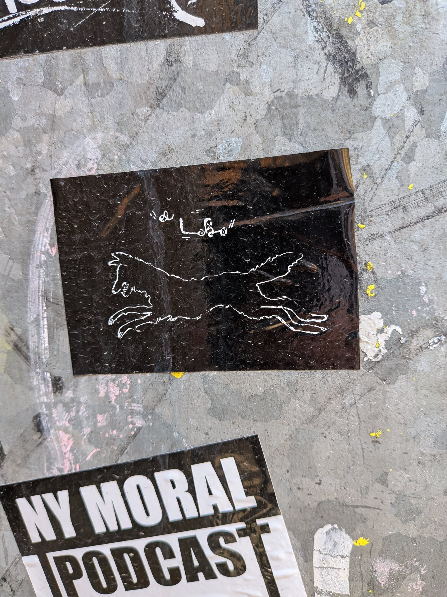 The sticker is a rectangular, glossy black background with a white outline drawing of a running animal, resembling a wolf or a wild dog, positioned centrally. The animal is depicted in mid-stride, with its head facing left and its mouth slightly open, showing teeth. Above the animal, in the upper left corner, white handwritten-style text reads 'la Lobo'. The sticker shows some wear, reflections, and minor creases, indicating it's affixed to a textured, possibly metallic, surface.