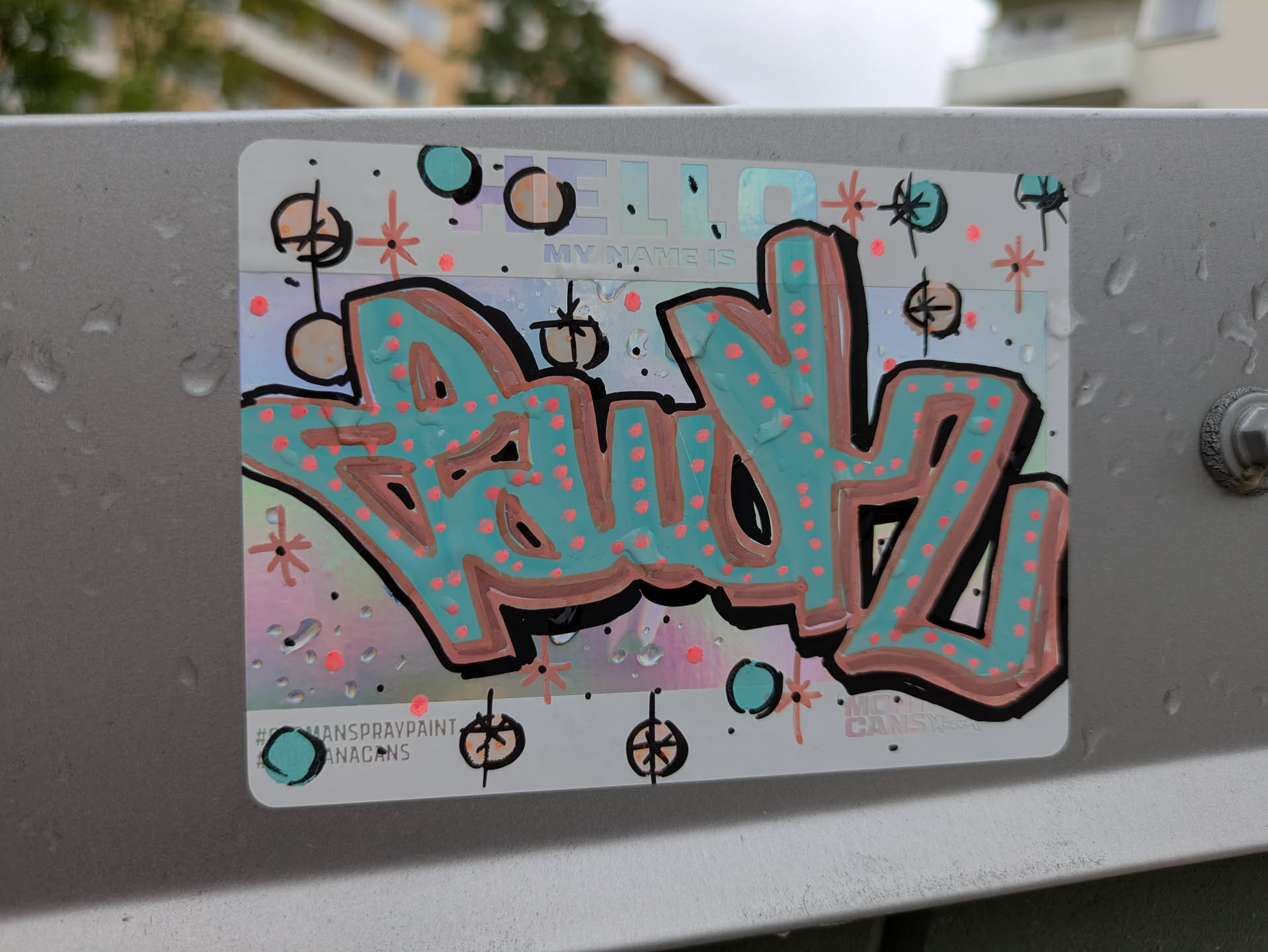 The sticker is rectangular with rounded corners, featuring a holographic background that reflects light with a rainbow sheen. The main element is a graffiti-style tag, 'BAUDZ', rendered in a prominent teal or light blue color with a thick black outline and a peach or light coral drop shadow. The teal letters are filled with numerous small, bright coral-pink dots. Above the tag, the faint, holographic text 'HELLO MY NAME IS' is visible. The background is further decorated with small, stylized starbursts in orange and black, and small teal and black circles, along with scattered black dots. Water droplets are visible on the sticker's surface, indicating it's outdoors. At the bottom left, there are two hashtags, and at the bottom right, additional text is present.
