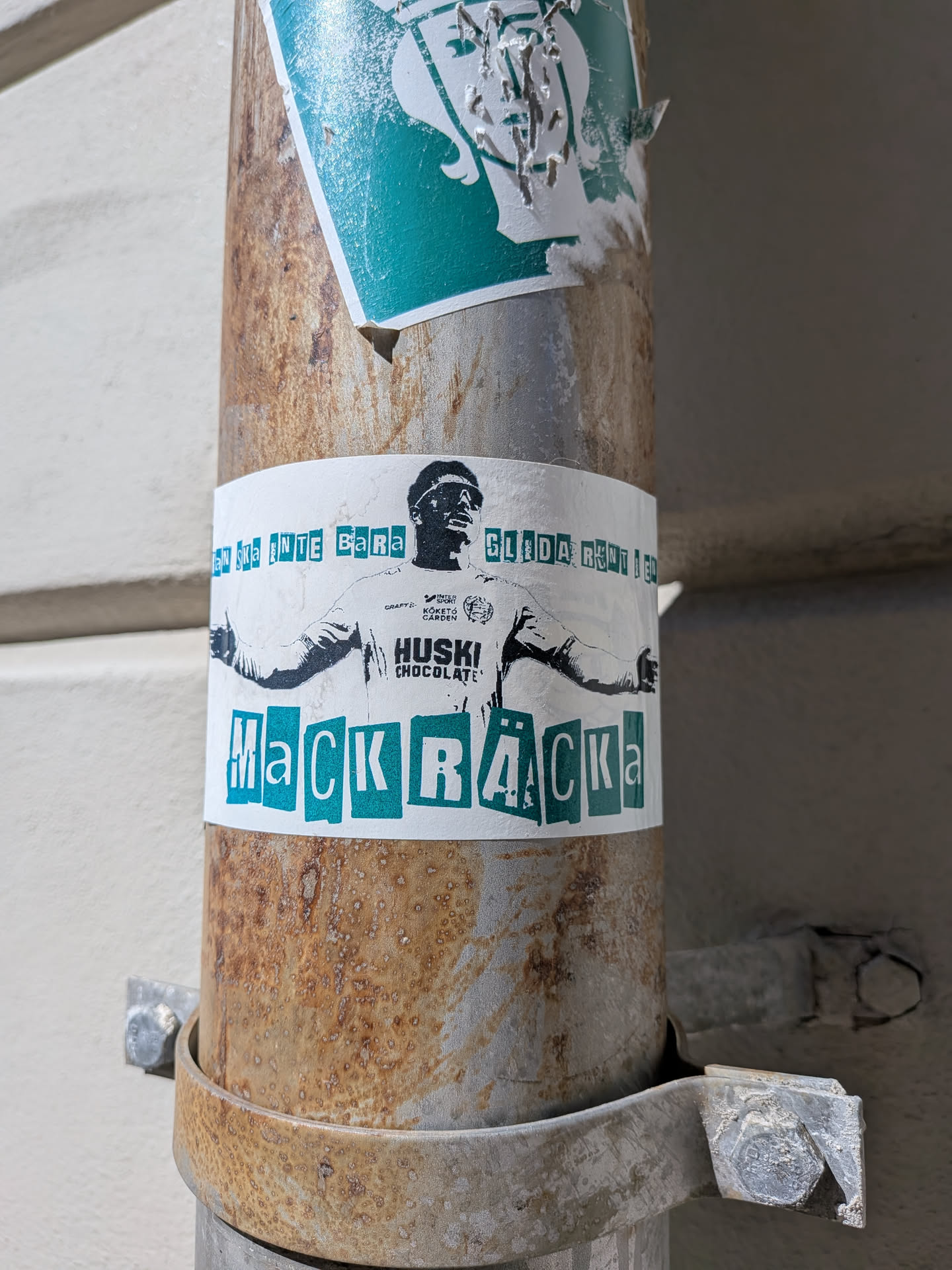 A rectangular white sticker featuring a black and white graphic illustration of a male figure, likely a football player, wearing sunglasses and a jersey with arms outstretched. The jersey displays several sponsor logos including 'CRAFT', 'INTERSPORT', 'KÖKETO GÅRDEN', and prominently 'HUSKI CHOCOLATE'. Above the figure, text in a distressed teal/turquoise color reads 'MAN SKA INTE BARA GLIDA RUNT I EN'. Below the figure, the word 'MACKRÄCKA' is spelled out in large, individually styled, teal/turquoise letters, giving a cut-out or stencil effect. The sticker is applied to a rusty metal pipe, showing some minor wear, dirt, and slight curvature.