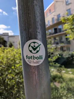 Street sticker A round white sticker on a metal pole. It displays a green soccer ball icon with a checkmark. Below, the green text 'fotbolli' is followed by Swedish text 'inga insatser - fina priser' and 'tippa på fotbolli.se' in black.