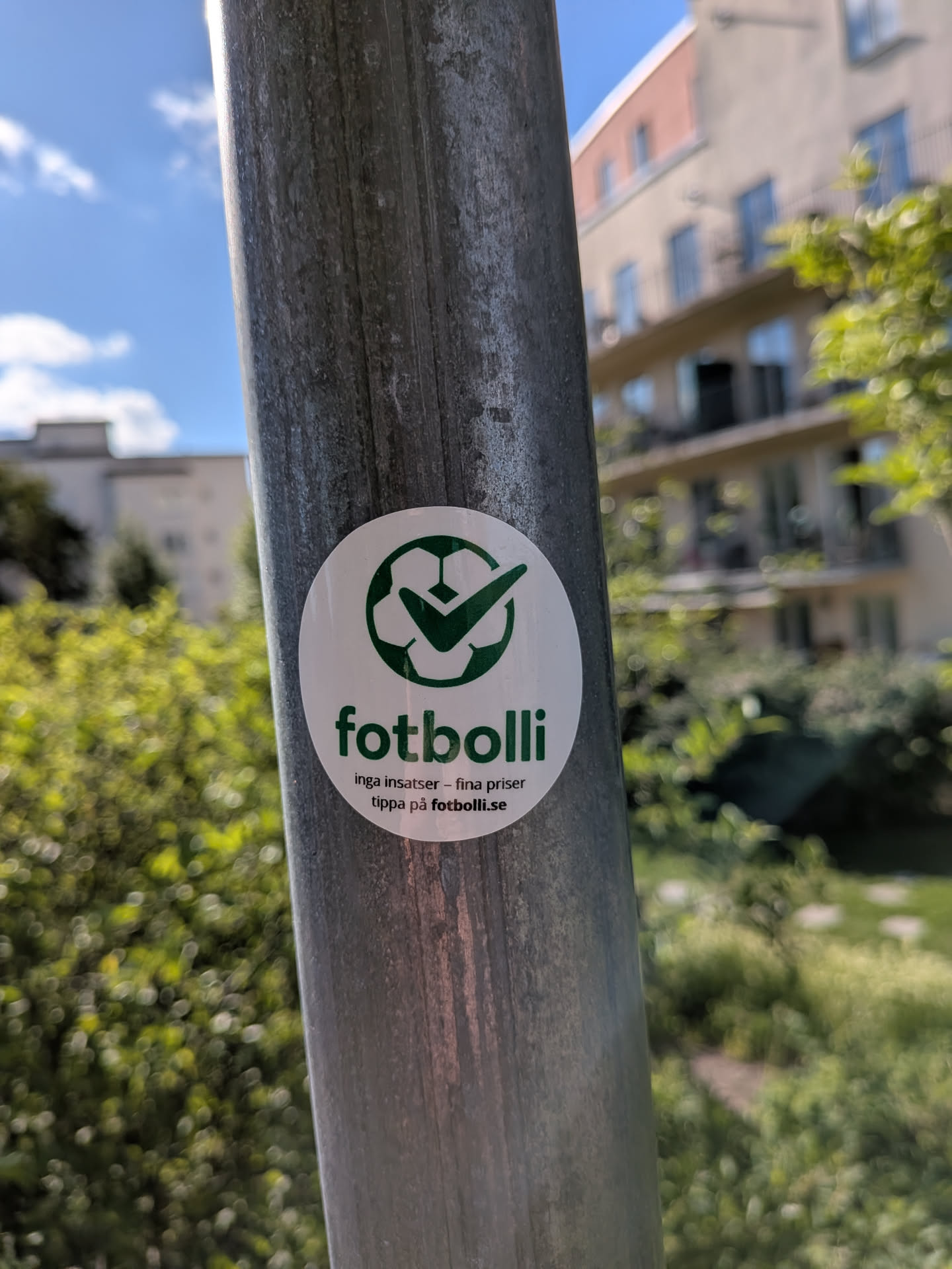 A round white sticker on a metal pole. It displays a green soccer ball icon with a checkmark. Below, the green text 'fotbolli' is followed by Swedish text 'inga insatser - fina priser' and 'tippa på fotbolli.se' in black.