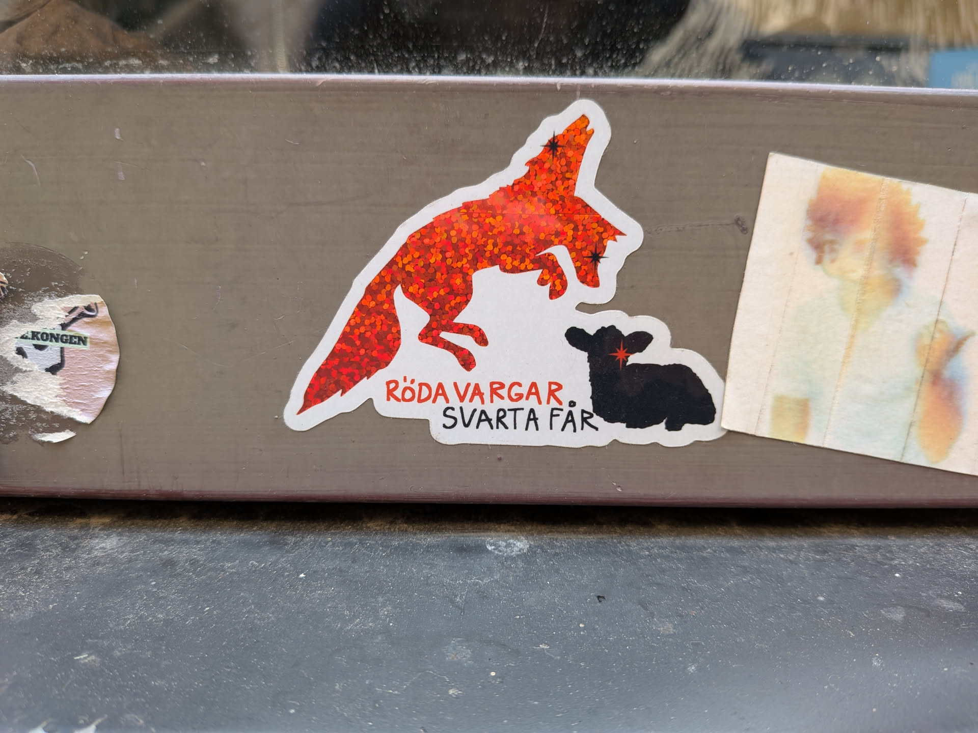 A die-cut sticker depicting a sparkling, red holographic wolf in mid-leap above a silhouette of a black sheep. Below them is the Swedish phrase 'RÖDA VARGAR, SVARTA FÅR' in red and black lettering. Both animals feature stylized star-shaped eyes.