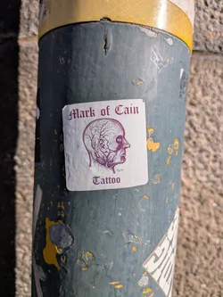 Street sticker The sticker is square with slightly rounded corners and has a white background. The prominent element is a detailed, profile illustration of a human head in deep purple ink. The illustration focuses on the vascular system of the head, showing numerous veins and arteries branching across the scalp and face. Below the head illustration, in a gothic-style font, reads 'Mark of Cain' in purple. Beneath that, in a smaller font, is 'Fig. 62.' and then 'Tattoo' in a similar gothic style.