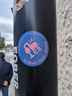 Street sticker A round sticker with a solid blue background. At the center is a pink illustration of a stylized four-legged creature whose neck curves downward, ending in a wine glass shape. Three small pink lines radiate above its back. Curving along the upper-left border is the text 'JUST SHARING LE BON MOMENT (NO ANIMALS WERE HARMED)' in small pink capital letters. Curving along the bottom-right border is the text 'DRINK FLUENT FRENCH' in larger pink capital letters, preceded by a pink Instagram camera icon.
