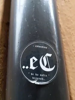 Street sticker A black and white circular sticker with the letters '.eC' in a gothic-style font.  Below it says "de ba sotla" bryppysh... in a smaller font. At the top, it says '- estocolmo -'. The sticker is applied to a dark gray cylindrical object.
