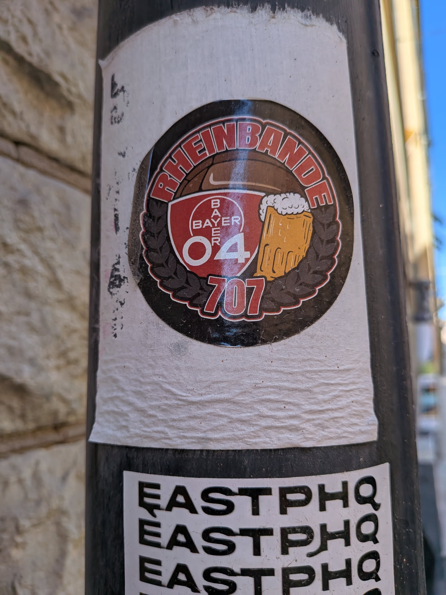 A circular sticker featuring a dark brown background resembling a football, complete with a white Nike swoosh. "RHEINBANDE" is written in red capital letters with a white outline along the top curve. Centered within the sticker is a red shield-like emblem displaying the 'BAYER' logo vertically, with '04' prominently in white with a red outline below it. To the right, a yellow beer mug with frothy white foam is depicted. Below these central elements, the number '707' is written in red with a white outline. The entire inner design is framed by a dark brown laurel wreath on either side. The sticker has a red and white outer border and is applied to a black pole, showing some signs of wear and minor scuffs.